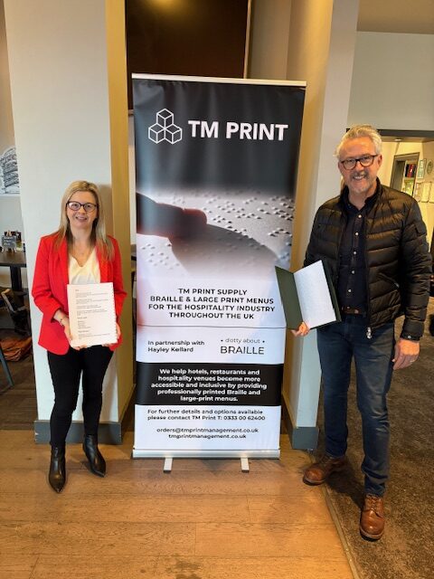 Hayley from Dotty About Braille stood with Glen from TM Print in front of a roller banner which advertises the braille and large print menus, they are each holding a menu.